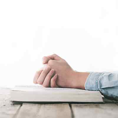 Two hands folded in prayer and resting on an open Bible.